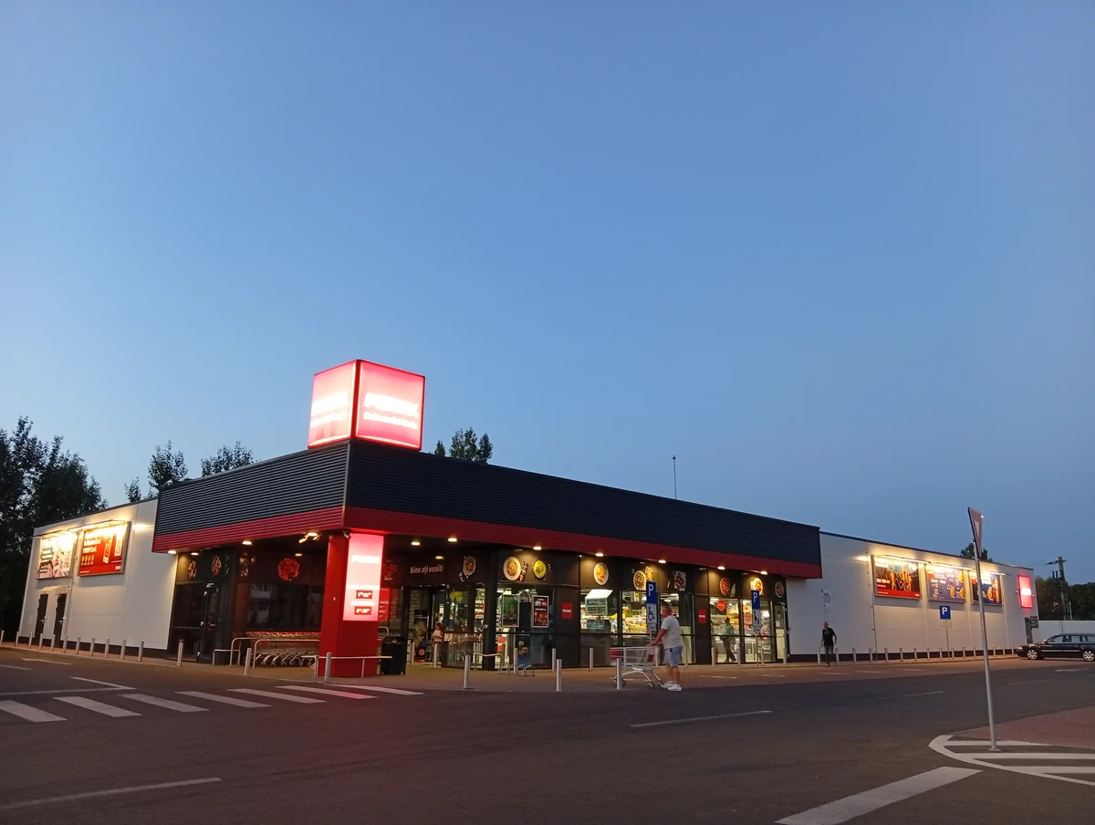 Penny supermarket in Craiova lit up at dusk, red sign glowing, a shopper with a trolley in front, 20 September 2026