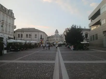 Piața Frații Buzești in Craiova at dusk — paved square, terraces with parasols on both sides, a domed building closing the view