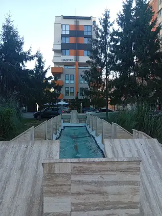 A stepped travertine water channel on Calea București in Craiova, jets running down a green-tiled bed towards the orange-and-grey Habitat building behind