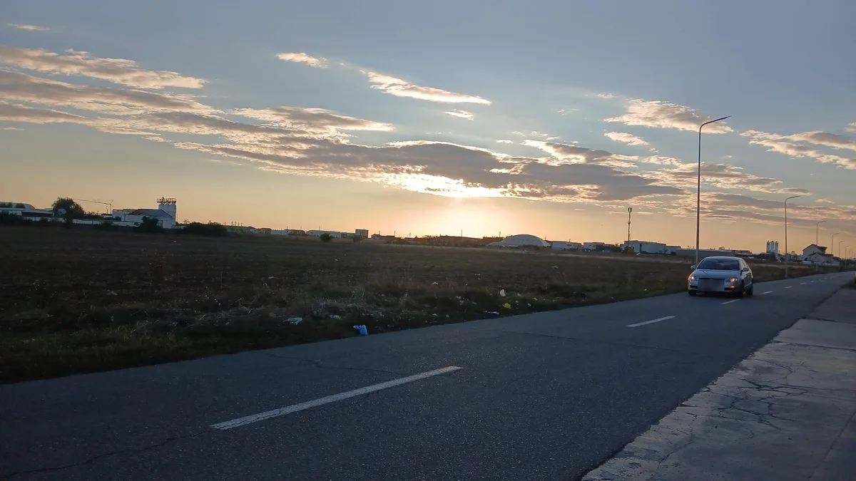 Sunset over a ploughed field on the industrial edge of the city, October 2026: warehouses, a silo and a white dome on the horizon, street lamps lining the road out of town