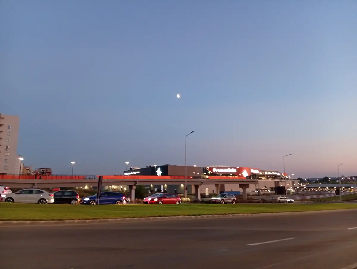 The mall from across the ring road at dusk, moon up over the Promenada sign — the view you get arriving from the north. September 2026