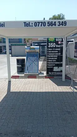 Open-air solar showroom in Craiova: white aluminium pergola with a stainless evacuated-tube water heater and a photovoltaic panel used as a price board beneath it